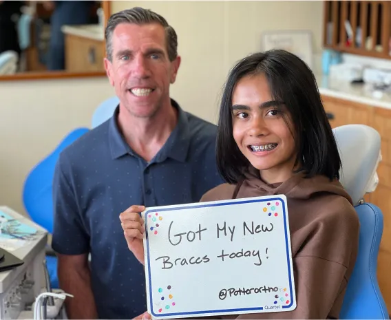 Orthodontist and patient celebrating new braces, girl holding sign "Got My New Braces today!" in dental office setting, showcasing orthodontic care at Potter Orthodontics.