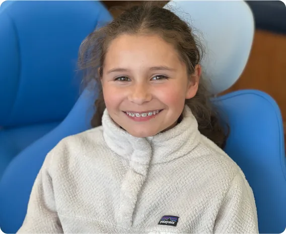 Smiling girl with braces wearing a cozy fleece jacket, seated in an orthodontic office chair, representing the journey of orthodontic treatment and the importance of maintaining a positive attitude while adapting to dietary changes.